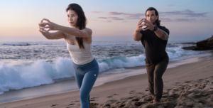 A man and woman practicing Foundation Training together on a serene beach, surrounded by gentle waves and a clear blue sky.