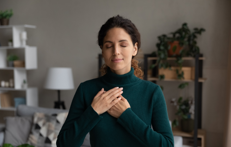 A woman with closed eyes and a peaceful expression places her hands over her heart. She is wearing a green turtleneck sweater, standing in a cozy, well-lit room with shelves, plants, and a lamp in the background.