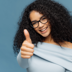 A young woman with curly hair and glasses smiles and gives a thumbs up, radiating positivity and confidence.