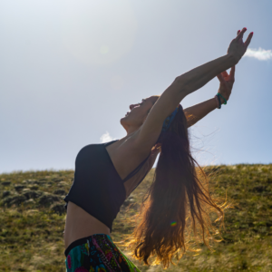 Person with long hair stretching their arms upward outdoors under a bright sky while being fully present.